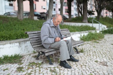 Yaşlı bir adam bankta otururken notlar alıyor, temiz havanın ve sessiz çevrenin tadını çıkarıyor.