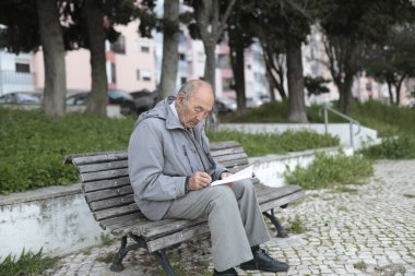 Parktaki bankta oturan yaşlı bir adam bir kağıt parçasına yazı yazıyor.