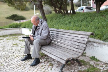 Yaşlı bir adam parktaki bankta otururken defterine not alıyor.