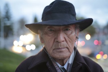 Şapka ve kulaklık takan yaşlı bir adamın portresi. Arka planda bulanık şehir ışıklarıyla kentsel bir ortamda duruyor.