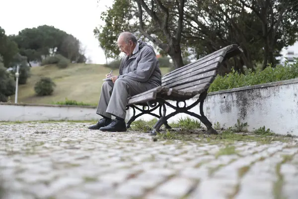 Huzurlu bir park ortamında ahşap bir bankta otururken not alan yaşlı bir adam.