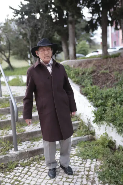 Şık bir kıdemli adam, şık bir şapka ve palto giyerek parkta rahatça yürüyor.