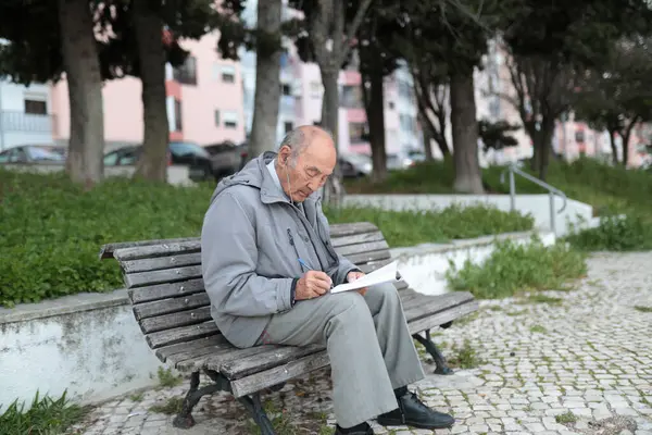 Parktaki bankta oturan yaşlı bir adam bir kağıt parçasına yazı yazıyor.