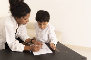 Oğluna matematik ödevinde yardım etmeye odaklanmış genç bir anne. Evdeki kağıdın üzerine kurşun kalemle numaralar yazıyor.