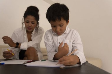 Genç çocuk sanatına konsantre olurken, renkli kalemlerle resim çizerken annesi de onun yaratıcı çabasını gözlemliyor ve destekliyor.