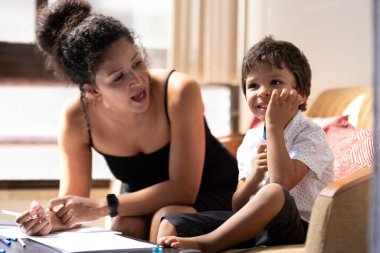 Bebek bakıcısı evde kanepede oturan gülümseyen bir anaokulu çocuğuna ev ödevlerini anlatıyor.
