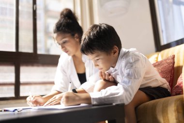 Odaklanmış genç bir çocuk ve annesi evde ödev yapıyorlar.