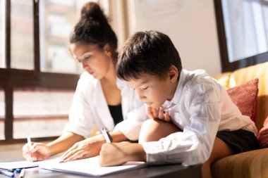 Çocuk ve anne kanepede oturuyor, deftere yazıyor, ödev yapıyorlar.