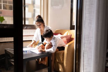 Anne, oğluna ev ödevinde yardım ediyor oturma odasında rehberlik ve destek sağlıyor.