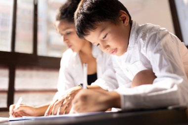 İlkokul öğrencisi, öğretmeniyle ya da evde eğitim gören annesiyle kağıda yazı yazıyor.