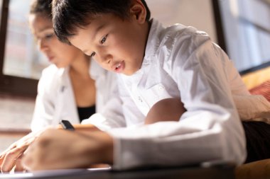 Asyalı etnik çocuk annesinin yanına resim çiziyor, ikisi de beyaz gömlek giyiyor ve bir kanepede oturuyor.