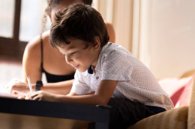 Küçük çocuk, annesinin yanına oturmuş ödevlerine yardım ediyor.