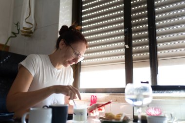 Yoğurt karıştıran ve kahvaltıda akıllı telefonunu kontrol eden bir kadın.