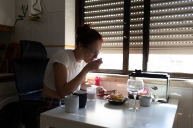 Modern bir mutfakta telefonunu kontrol eden bir kadın kahvaltıdan zevk alıyor.