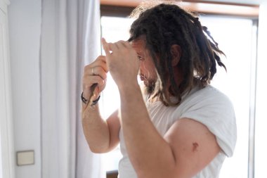 Kafkas saçlı bir adam sabahları saçını şekillendiriyor, pencerenin yanındaki bir yatak odasında duruyor.