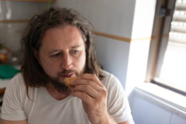 Uzun saçlı ve sakallı beyaz bir adam evcil bir mutfakta kurabiye yiyor.