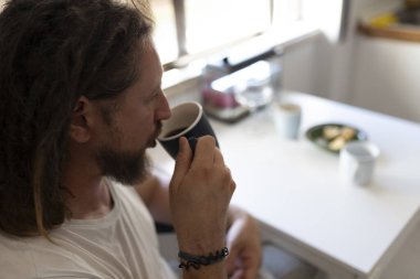 Beyaz bir adam evinde, mutfağında bir fincan kahvenin keyfini çıkarıyor.