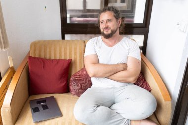 Kendine güvenen erkek serbest çalışan, dizüstü bilgisayarın yanındaki koltukta çapraz kollarıyla oturuyor, uzaktan çalışmanın tadını çıkarıyor ve başka tarafa bakıyor.