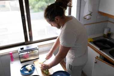 Bir adam, küçük bir teraryumun yanında, mutfaktaki ahşap bir tahtada sebze keserek yemek hazırlıyor.