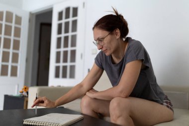 Oturma odasındaki kanepede otururken not defterine notlar yazan gözlüklü olgun bir kadın.