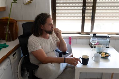 Rastalı bir adam iş molasında ev mutfağında kahvaltı edip kahve içiyor.