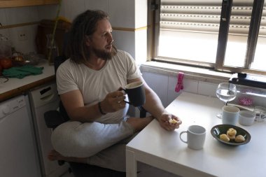 Uzun saçlı ve sakallı genç adam evden çalışırken kahvaltının ve kahvenin tadını çıkarıyor.