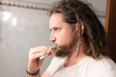 Rastalı beyaz bir adam diş fırçasıyla dişlerini fırçalıyor, tuvalette ağız sağlığını koruyor.