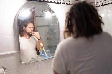 Kafkasyalı bir adam banyodaki aynada dişlerini fırçalıyor, günlük rutinini yansıtıyor.