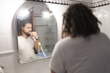 Banyo aynasında dişlerini fırçalayan sakallı ve rastalı bir adam günlük ağız temizliğini yansıtıyor.