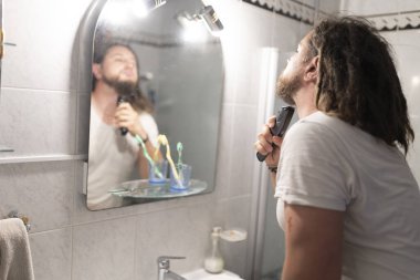 Rastalı bir adam banyodaki aynanın önünde sakalını tıraş ediyor.