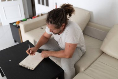 Evdeki rahat kanepeye oturmuş not defterine notlar yazan konsantre bir adam.