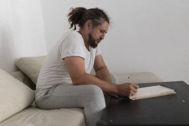 Evdeki kanepede otururken not defterine odaklanmış bir adam.