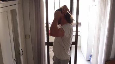 Uzun saçlı bir adam saçlarını topuz şeklinde bağlıyor, pencerenin yanında.