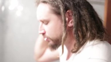 Rastalı beyaz bir adam yüzünü banyo lavabosunda yıkıyor.