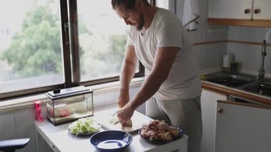 Genç adam küçük mutfağında tahta bir tahtanın üzerinde tavuk bacağı kesiyor.