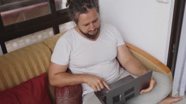 Rastalı ve sakallı genç adam oturma odasındaki kanepede otururken dizüstü bilgisayar kullanıyor.