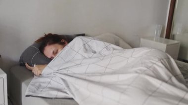 Uykulu kadın yatakta uzanıyor, battaniyenin altına saklanıyor ve esniyor.