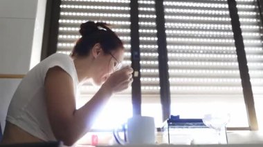 Genç bir kadın kahvaltı yapıyor ve pencerenin yanındaki telefonuna bakıyor.