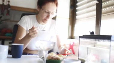 Genç bir kadın kahvaltısını evde yapıyor, akıllı telefonunu kontrol ediyor ve akvaryuma bakıyor.