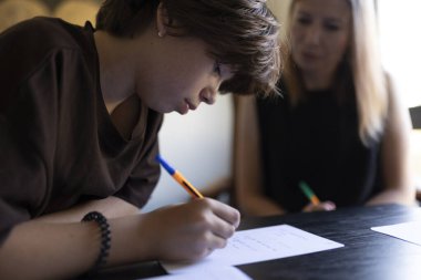 Odaklanmış ergen öğrenci sınıfta notlar yazarken öğretmen asistanlık yapıyor.