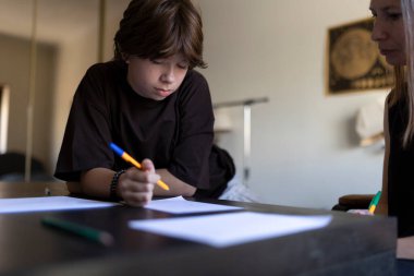 Okul sonrası özel ders sırasında bir ev öğretmeni tarafından yönetilen, kağıda yazı yazan genç bir öğrenci.