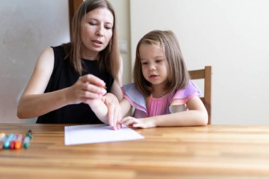 Küçük bir kıza ahşap bir masada yazı yazması için yardım eden evde eğitim öğretmeni.
