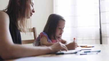 Genç anne, küçük kızına evde keçeli kalemlerle resim çizmesinde ve resim çizmesinde yardım ediyor.
