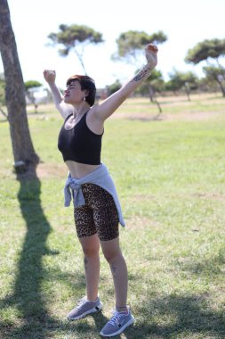 Parkta güneşli bir günün tadını çıkaran genç bir kadın. Kollarını açıp antrenmana hazırlanıyor.
