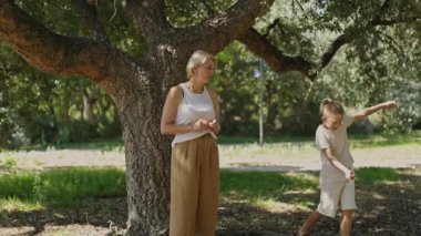 Anne ve çocuklar parkta bir ağacın altında oynayıp eğleniyorlar.