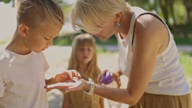 Bakıcı anne, güneşli bir parkta oğullarına dezenfektan sürüyor. Hijyen ve güvenliği sağlıyor.