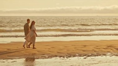 Genç çift gün batımında sahilde romantik bir yürüyüş yapıyor.