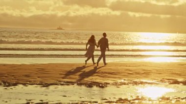 Genç çift gün batımında sahilde romantik bir yürüyüş yapıyor.