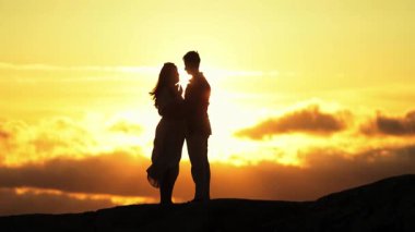 Sevgi dolu bir çiftin silueti bir tepenin üstünde canlı bir günbatımı arkaplanına sarılıyor.