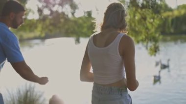 Güneşli bir günde göl kenarında kazları besleyen mutlu bir aile.